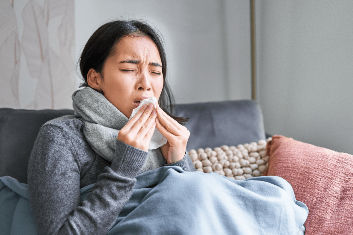 A woman at home, wrapped in warm clothes and a scarf, looking unwell and experiencing symptoms of a Cough